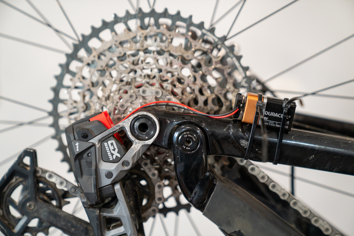 Close-up of a bicycle's rear derailleur and cassette, featuring a SRAM GX derailleur mounted on the frame. A 9V battery is secured with zip ties, connected to the derailleur, highlighting the electronic shifting mechanism. The background includes bicycle spokes and part of the cassette.