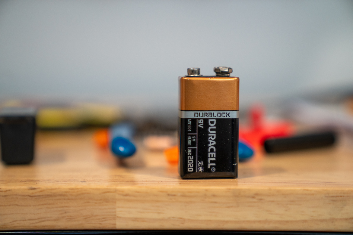 A close-up view of a 9-volt Duracell battery placed on a wooden surface, with various other small objects and tools slightly out of focus in the background. The battery features a black and gold design with labels indicating its brand and specifications.
