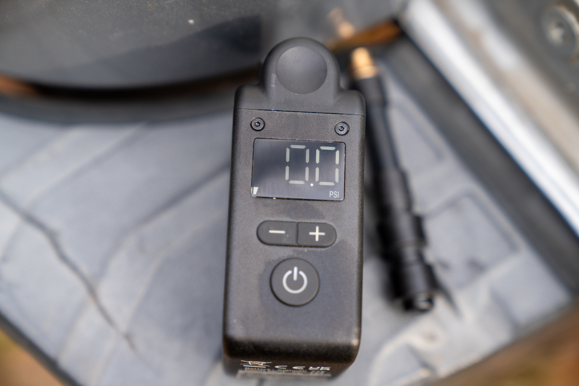 Digital tire pressure gauge displaying a reading of 0.0 PSI, with buttons for power and adjusting pressure. An air hose connector is visible, resting on a textured surface.