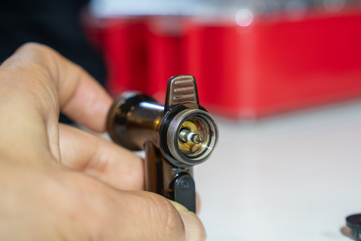 A close-up image of a person holding a brass-colored air brush tool with a visible nozzle, set against a blurred background with red and gray elements. The focus is on the mechanism of the air brush, highlighting its detailed design and part of the user's hand.