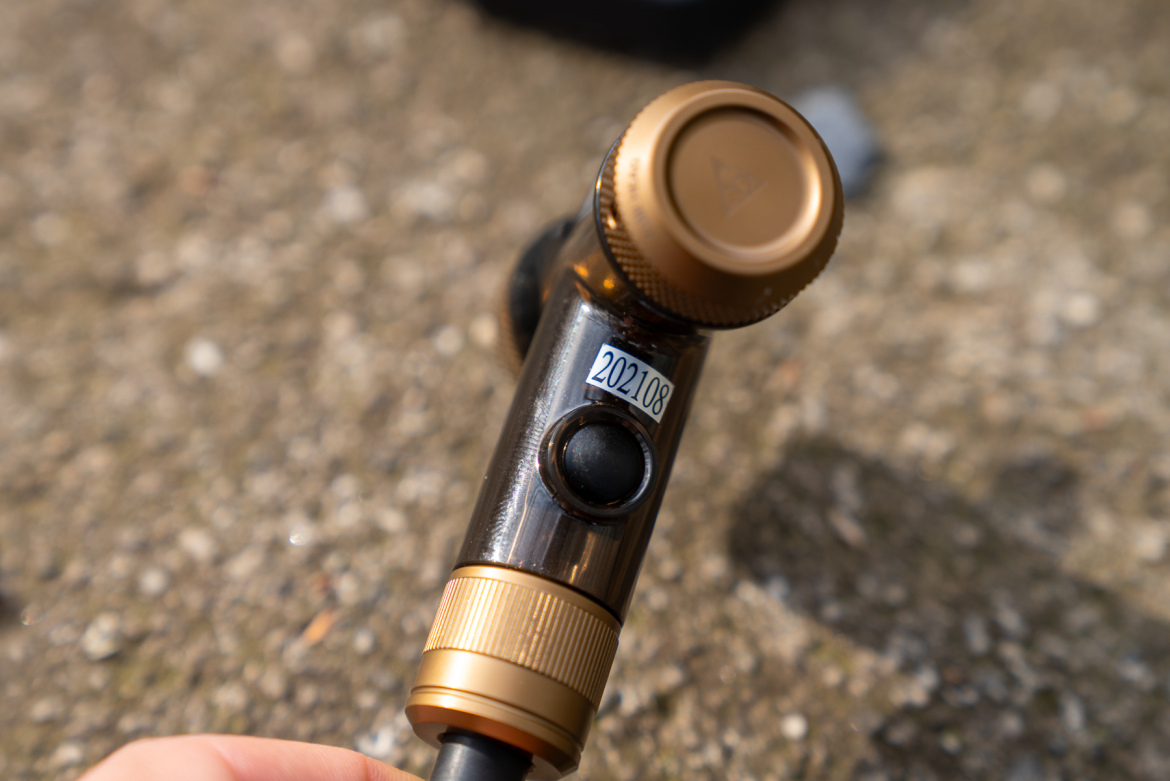 Close-up image of a dark, metallic device with a gold-colored knob and button, resting on a textured surface. The device has a label with the numbers "202108" visible.