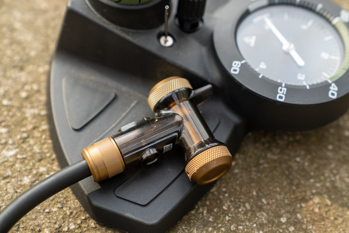 Image of a close-up view of a black dive computer with a pressure gauge, featuring a brass-colored air connector attached to a rubber hose. The background shows a textured surface.