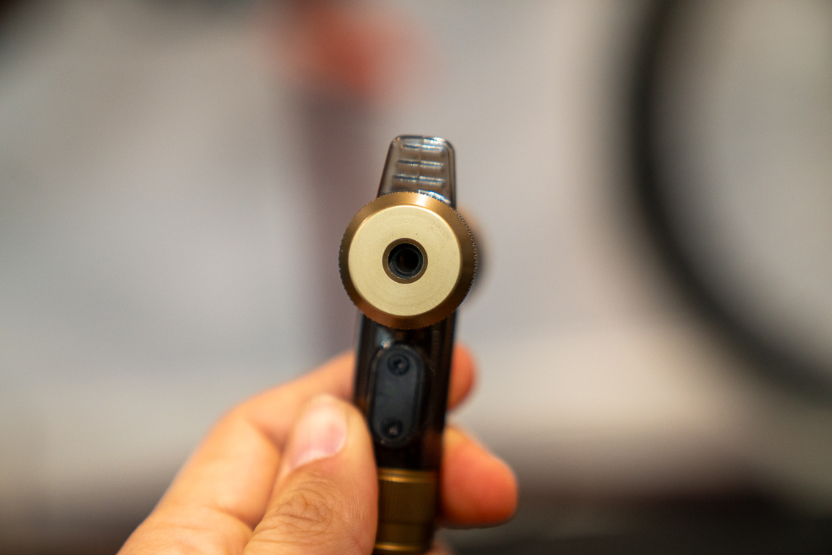 A close-up image of a brass-colored valve with a circular opening, held in a person's hand. The background is blurred, suggesting a workshop or indoor setting.