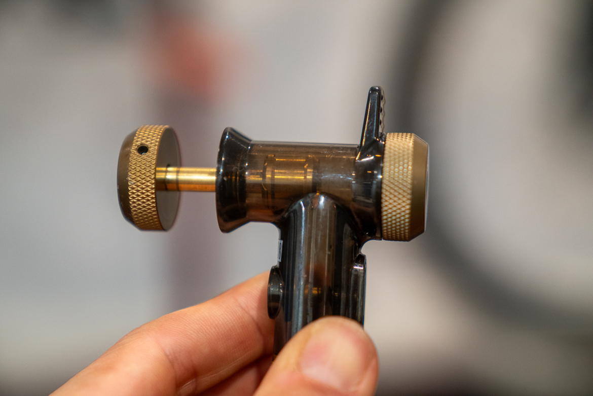 Close-up image of a hand holding a small mechanical tool with a black and gold design, featuring a knob and nozzle. The tool is likely used for precise applications, possibly in repair or manufacturing. The background is blurred, focusing on the tool's details.
