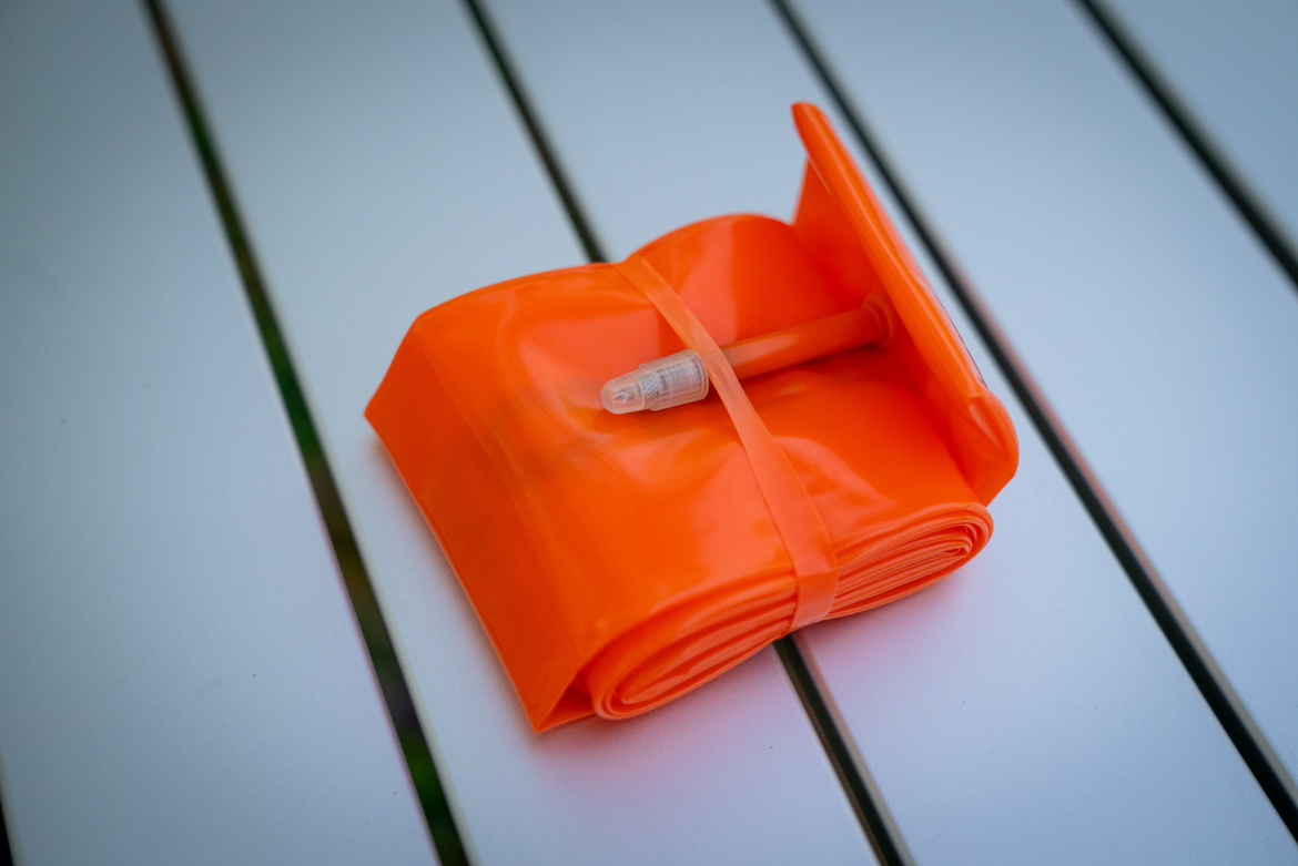A rolled-up orange flexible strip with a small connector, resting on a white surface with parallel lines. The strip is secured with a band, and it appears to be made of a glossy material.