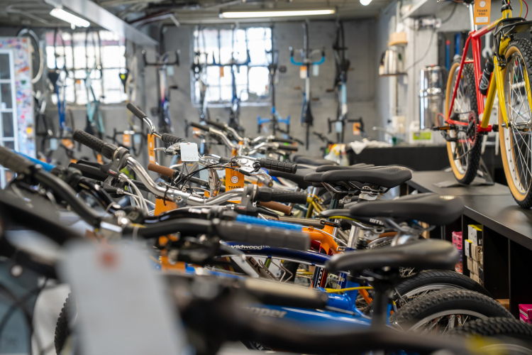Interior view of a bike shop featuring a variety of bicycles on display. In the foreground, several bikes with different colored frames are lined up, showcasing their handlebars and seats. In the background, more bicycles can be seen hanging on the wall. The shop has a modern feel with organized shelves and equipment visible.