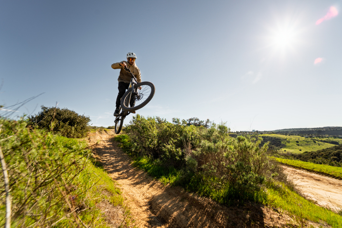 A mountain biker performing a jump on a dirt trail surrounded by greenery, under a clear blue sky with a bright sun shining in the background.