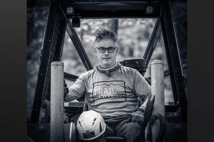 A man sitting in the driver's seat of a tractor, wearing a helmet and a shirt that reads "Trail Dynamics." He has glasses and is smiling at the camera. The image is in black and white, with blurred greenery in the background.