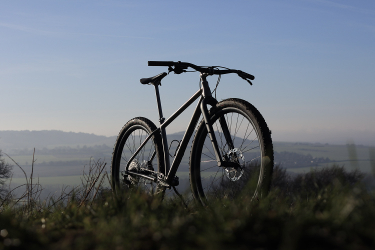 A mountain bike is displayed on a grassy hilltop with a scenic background of rolling hills under a clear blue sky. The bike is positioned in silhouette, showcasing its frame and tires, with a focus on the rugged terrain and distant landscapes.