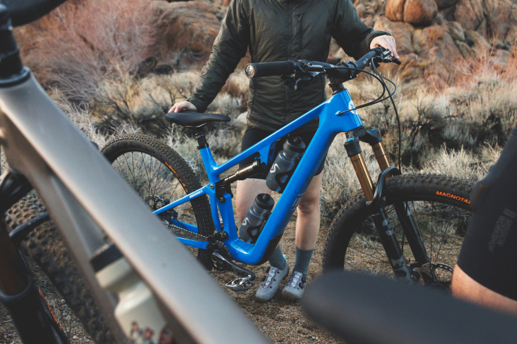A person in a green jacket stands next to a blue mountain bike with two water bottles mounted on the frame. The background features rocky terrain and sparse vegetation, indicating an outdoor setting suitable for biking. The person wears shorts and gray socks with shoes, suggesting they are prepared for a biking adventure.