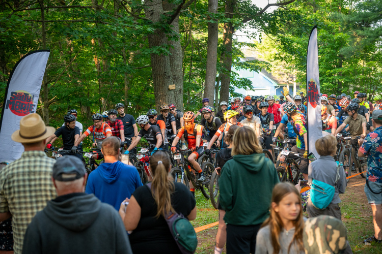A crowd of mountain bikers is gathered for a race in a wooded area. They are lined up on their bicycles, wearing helmets and cycling gear, with a few spectators in the foreground. A banner with the event's logo is visible, and the scene is set in a lush green environment, suggesting a vibrant outdoor atmosphere.