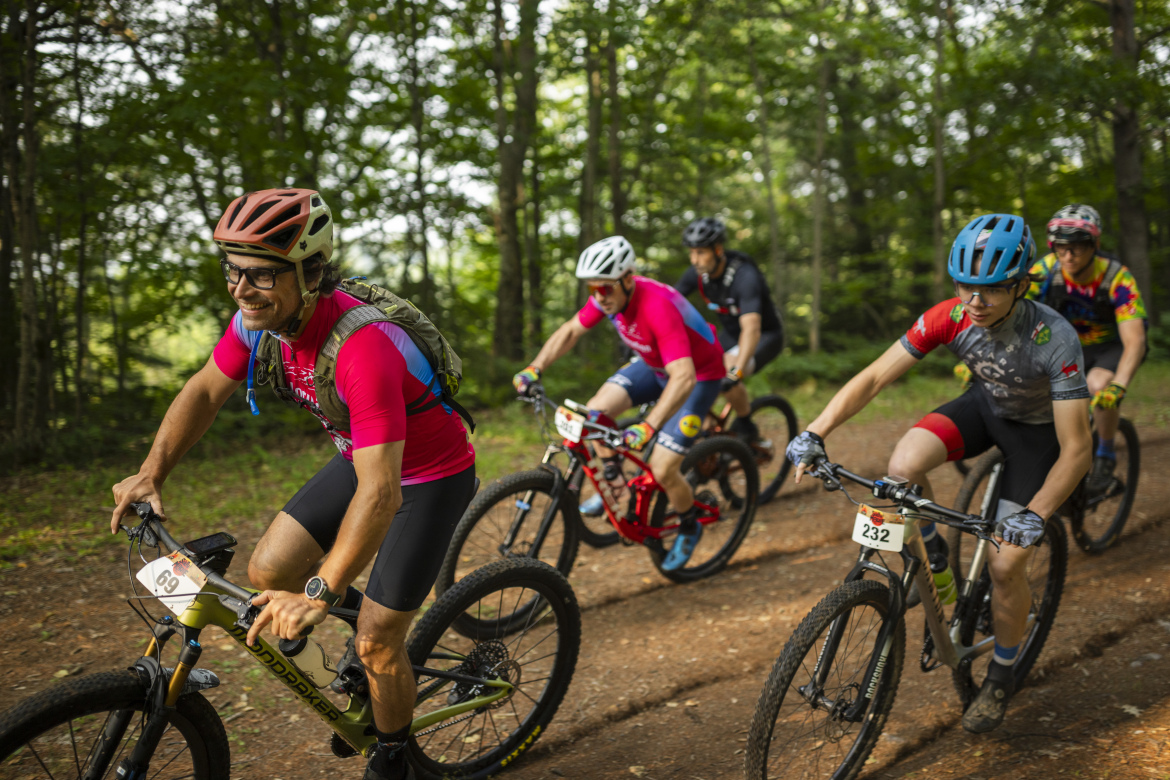 A group of five male mountain bikers riding on a dirt trail through a forested area. The lead rider, wearing glasses and a helmet, is smiling while in a bright pink shirt and black shorts. The other cyclists are dressed in various colorful jerseys and helmets, with two sporting numbers on their bikes. The atmosphere is energetic and lively, surrounded by green trees and natural scenery.
