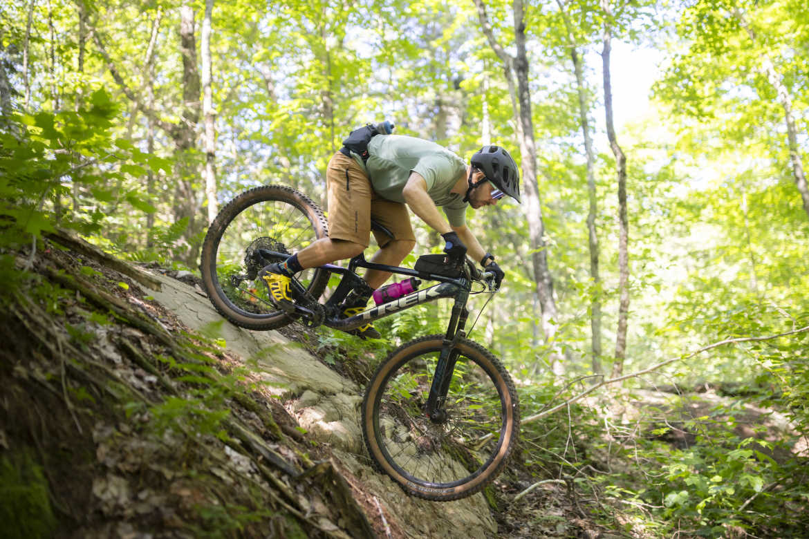 A mountain biker riding down a rocky slope in a lush green forest, wearing a helmet, gloves, and casual clothing. The bike is equipped for off-road terrain, and the cyclist leans forward to maintain balance as they navigate the challenging trail surrounded by trees and foliage.