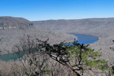 A panoramic view of a winding river surrounded by barren hills and trees under a clear blue sky. The landscape features rugged cliffs on one side and gentle slopes on the other, showcasing the natural beauty of the area. Raccoon Mountain Trail Network mountain bike trail.