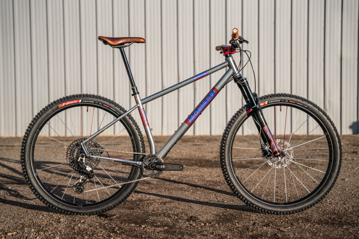 A modern mountain bike with a sleek silver frame, featuring a brown leather saddle and prominent knobby tires, positioned on gravel with a corrugated metal backdrop.