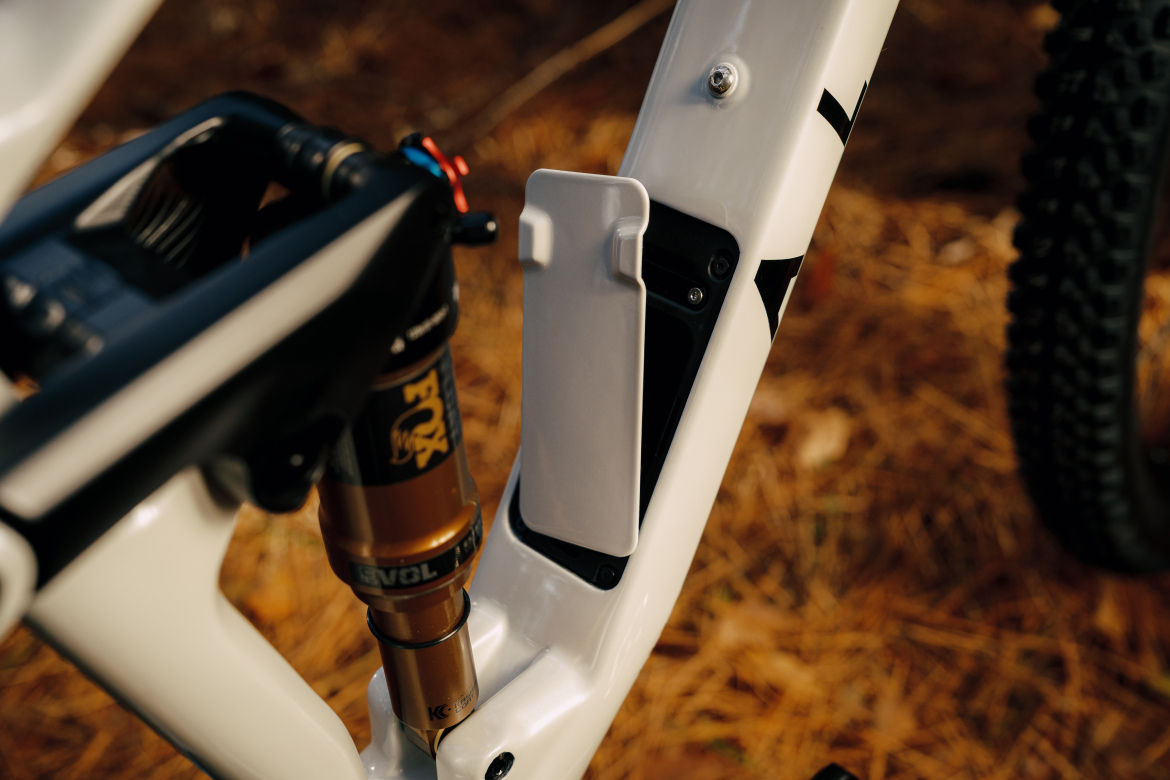 Close-up of a white mountain bike frame showcasing the shock absorber and a storage compartment integrated into the frame, with a backdrop of pine needles. The image highlights the bike's sleek design and engineering features.