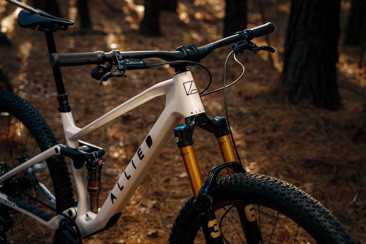 Close-up of a modern mountain bike in a forest setting, featuring a white frame with the brand name "ALLIED" displayed prominently. The bike's front suspension showcases gold forks, and the handlebars are equipped with grips and brake levers. Sunlight filters through the trees, illuminating the bike and highlighting the natural surroundings, which include a forest floor covered in pine needles.