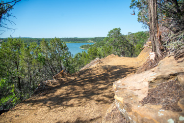 A scenic trail overlooking a lake, framed by greenery and rocky outcrops. The path appears well-maintained, winding along the edge of a cliff with clear blue skies above. Palo Pinto Mountains State Park mountain bike trail.