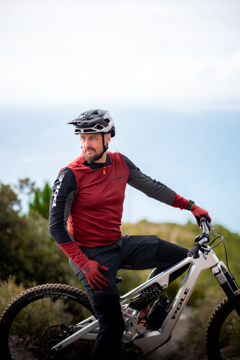 Leo Kokkonen wearing a red and black cycling jersey, black pants, and a helmet sits on a white mountain bike, looking thoughtfully into the distance. He is outdoors with a soft-focus background of greenery and the ocean visible in the distance, conveying a sense of adventure and the thrill of mountain biking.