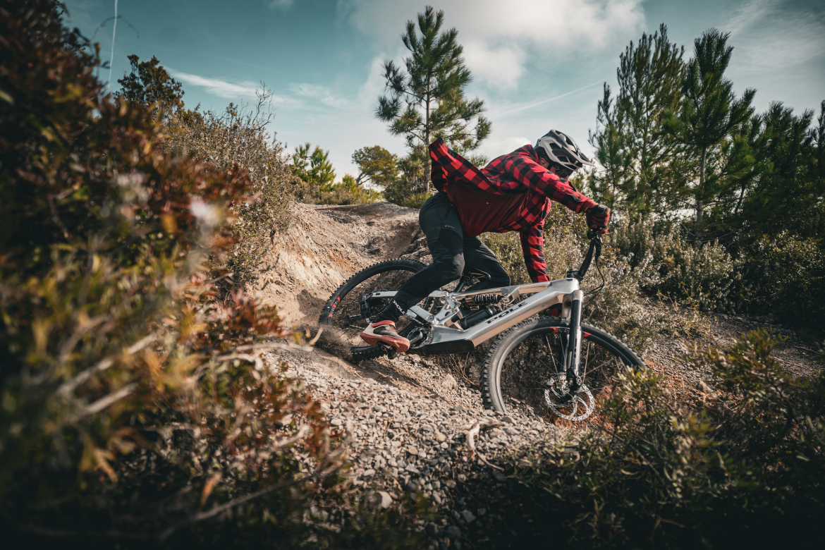 A mountain biker wearing a red and black flannel shirt and a helmet is navigating a rocky trail surrounded by greenery. The bike is in motion, with one tire elevated as the rider leans into a turn. The sky is partly cloudy, contributing to the adventurous outdoor atmosphere.