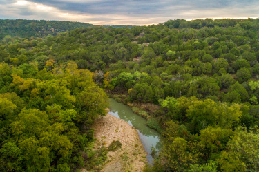 Aerial view of a winding river surrounded by lush greenery and rolling hills, with a cloudy sky above, showcasing the beauty of a natural landscape. Palo Pinto Mountains State Park mountain bike trail.