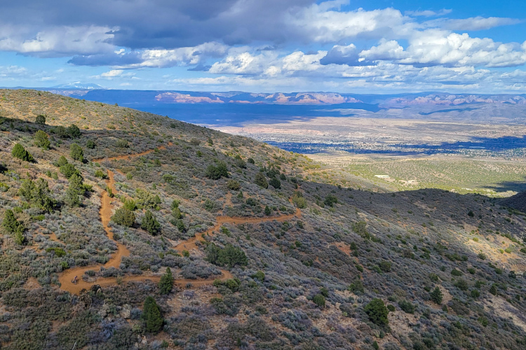 A scenic view of rolling hills covered with shrubs and trees, featuring a winding dirt path. In the background, expansive valleys and distant mountains are visible under a partly cloudy sky. The landscape showcases a mix of greenery and earth tones, exemplifying a natural outdoor setting.