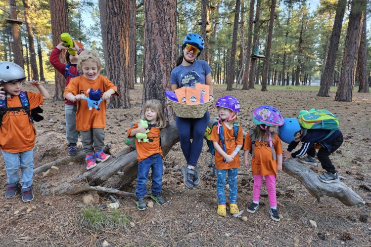 A group of young children wearing orange shirts and various helmets are playing in a forest. Some are standing on a fallen log, while one child is holding a toy and another has a small backpack. An adult, also in casual clothing, is smiling and holding a basket filled with items. The scene captures a joyful outdoor activity among tall trees and pine needles.