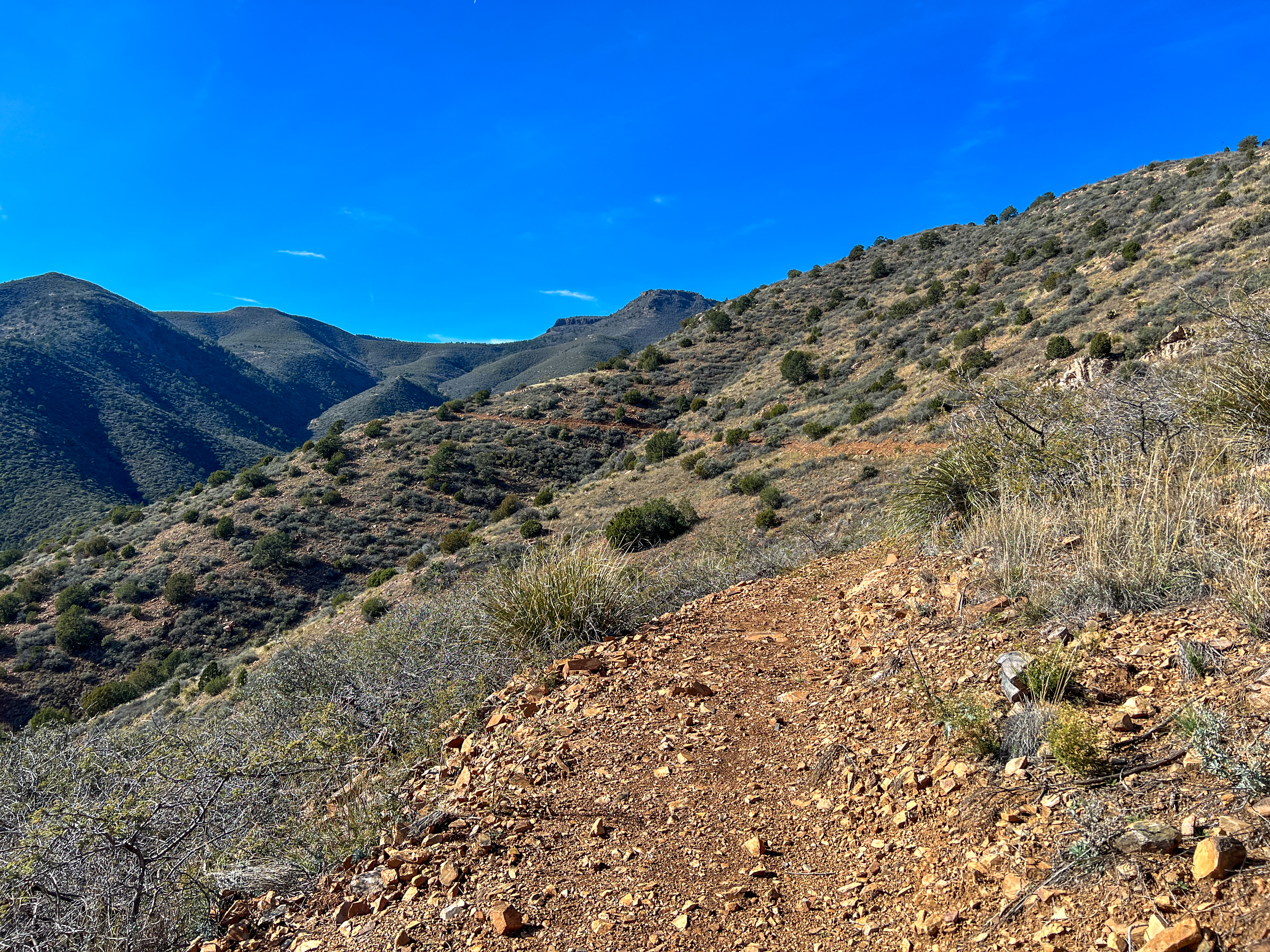A winding dirt path leads through a mountainous landscape, surrounded by shrubs and sparse vegetation. The hills, covered in green and brown, rise gently towards a clear blue sky, showcasing a serene outdoor setting perfect for hiking. Copper Chief mountain bike trail.