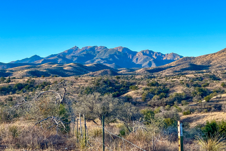 A scenic view of mountains under a clear blue sky, featuring rolling hills and sparse vegetation in the foreground, with a faded fence line adding depth to the landscape.