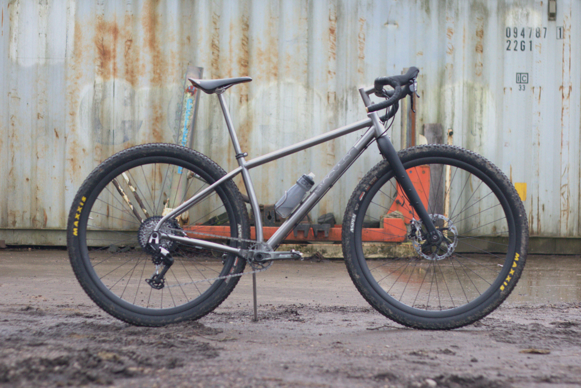 A modern mountain bike with a titanium frame, wide tires, and disc brakes, positioned in front of a weathered metal container. The bike features a water bottle mounted on the frame and is shown on a muddy surface, highlighting its rugged design.