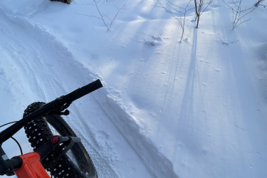 A close-up view of a fat tire bike riding on a snowy trail in a forest. The image captures the front wheel and part of the rider's foot positioned on the pedal, with snow-covered logs and trees in the background, illuminated by soft sunlight. Mud Pond Loop mountain bike trail.