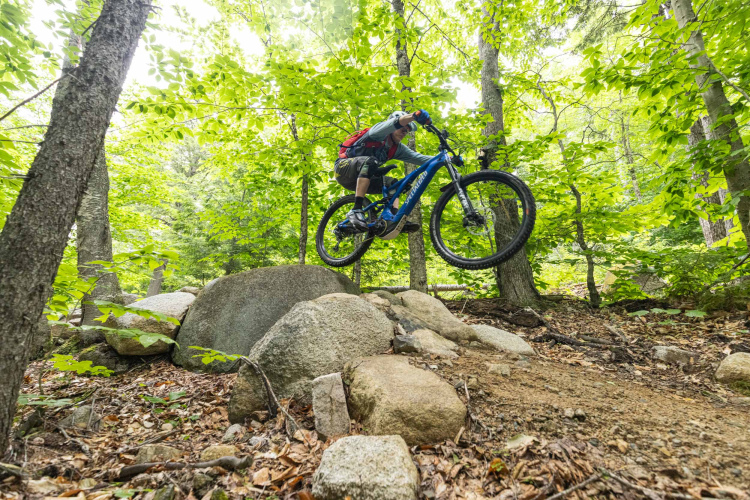 A mountain biker performing a jump over a large rock on a forest trail, surrounded by vibrant green trees and foliage.
