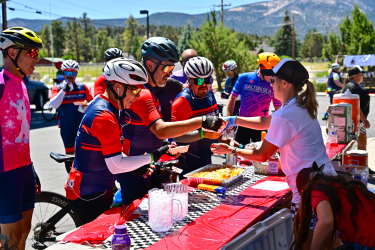 A group of cyclists in bright jerseys and helmets gather around a red table at an outdoor event, receiving food and drinks from a volunteer. The scene is set against a backdrop of trees and mountains under a sunny sky. Several cyclists are holding plates and cups, while others chat and share moments of camaraderie.