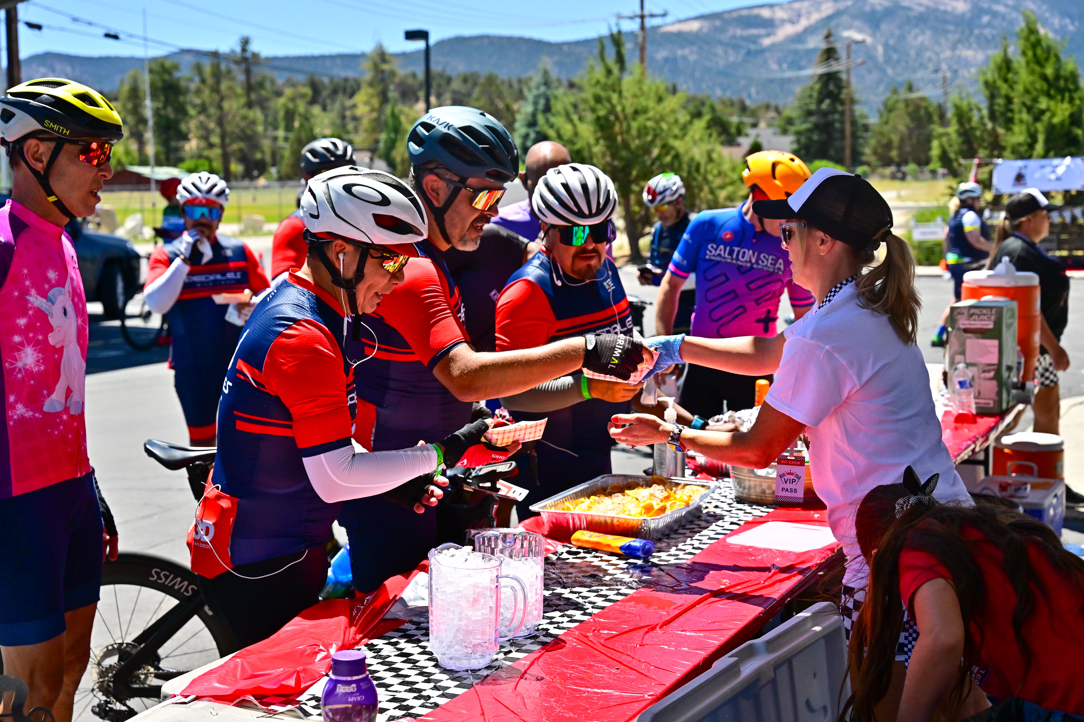 A group of cyclists in bright jerseys and helmets gather around a red table at an outdoor event, receiving food and drinks from a volunteer. The scene is set against a backdrop of trees and mountains under a sunny sky. Several cyclists are holding plates and cups, while others chat and share moments of camaraderie.