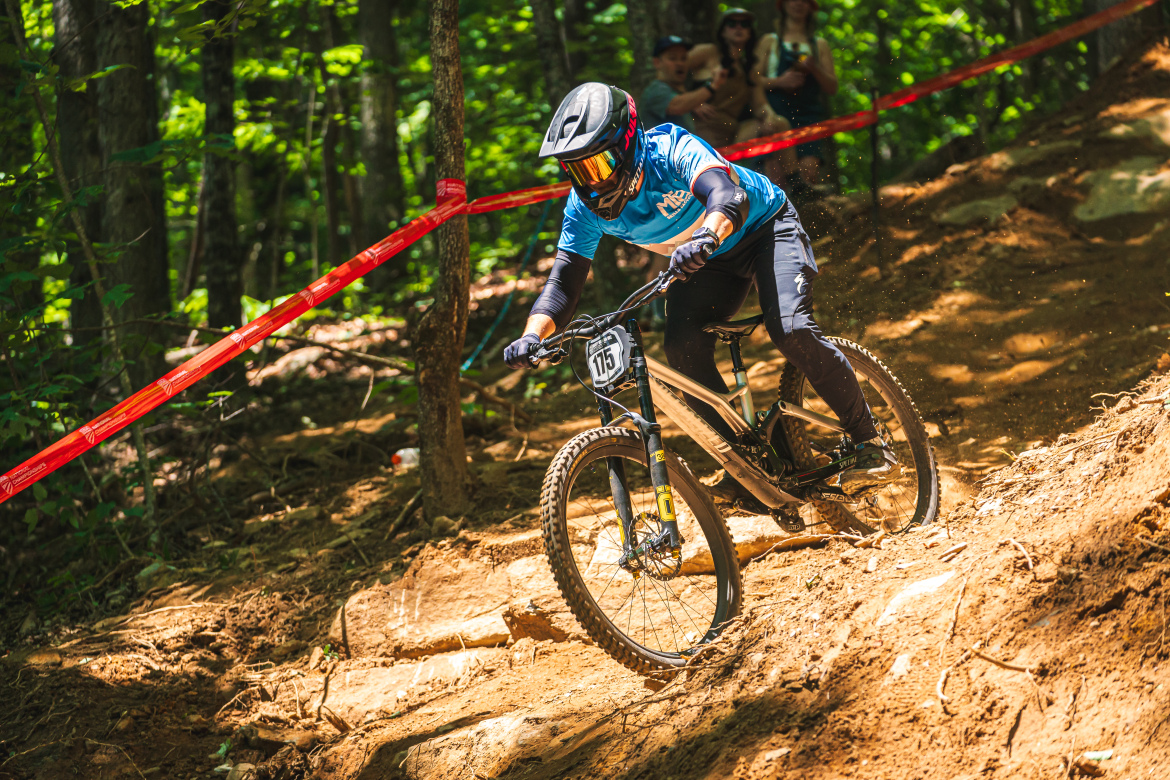 A mountain biker skillfully navigating a downhill trail in a forested area, surrounded by trees and marked with red tape. The rider is wearing a helmet and protective gear, focused on maintaining balance while riding on uneven terrain. In the background, spectators can be seen watching the race.