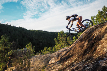 A mountain biker skillfully navigating a rocky terrain, leaning forward as they ride down a steep rock face amidst a lush green forest under a bright blue sky with wispy clouds. Santa Coloma de Farners mountain bike trail.