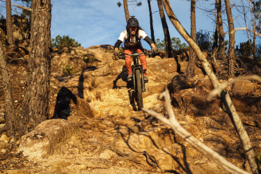 A mountain biker navigating a rocky trail surrounded by trees on a sunny day. The rider is airborne, with the bike in mid-jump as they approach a steep descent. Santa Coloma de Farners mountain bike trail.