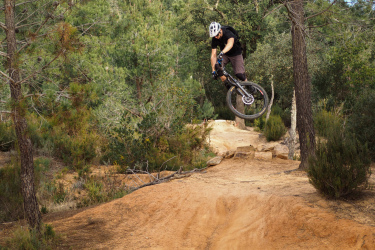 A mountain biker in mid-air performing a jump over a dirt ramp in a forested area, surrounded by trees and vegetation. Santa Coloma de Farners mountain bike trail.