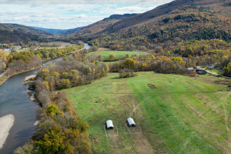Aerial view of a serene landscape featuring a winding river surrounded by lush green fields and rolling hills. The foreground includes two metallic structures on the grass, while a backdrop of autumn-colored trees decorates the hillsides. In the distance, a small town is visible along the riverbank, set against a clear blue sky with scattered clouds.