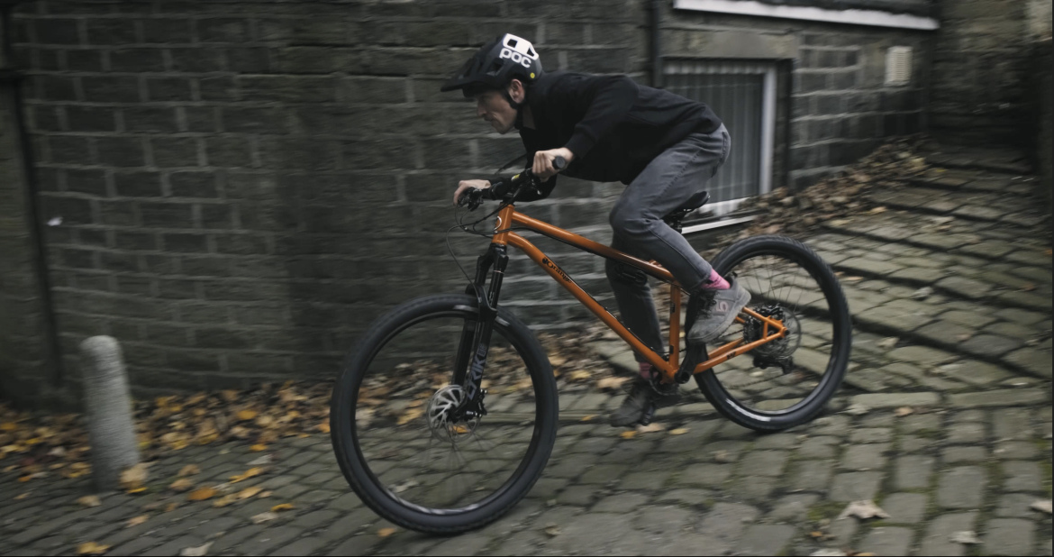 A person riding an orange mountain bike at speed on a cobblestone path, wearing a black helmet and dark clothing, with fallen leaves scattered on the ground and brick walls in the background.