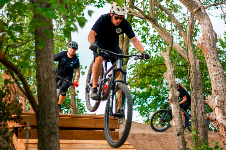 Two mountain bikers navigating a wooden ramp in a forested area. One cyclist is airborne, while the other is riding closely behind on the ground. Lush greenery surrounds them, with trees framing the scene. The atmosphere conveys excitement and action in a natural setting.