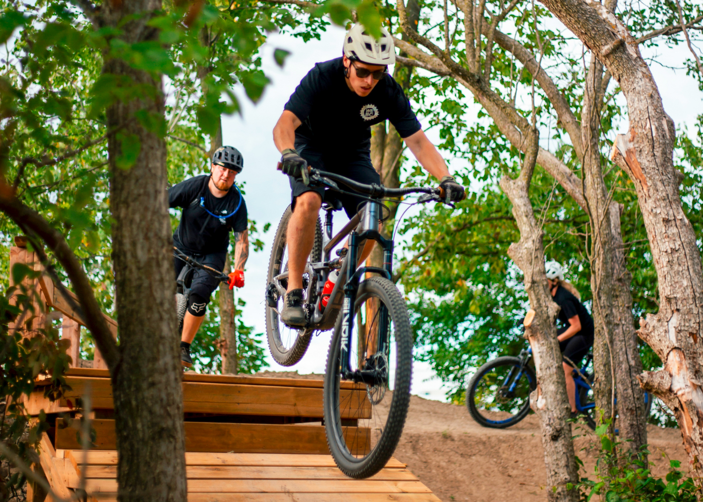 Two mountain bikers are navigating a wooden ramp in a forested area. One rider is airborne, performing a jump, while the second is following closely behind on the ramp. The backdrop features trees and greenery, indicating an outdoor biking trail. Both riders are dressed in appropriate biking gear, including helmets. Libro Centre Trails mountain bike trail.