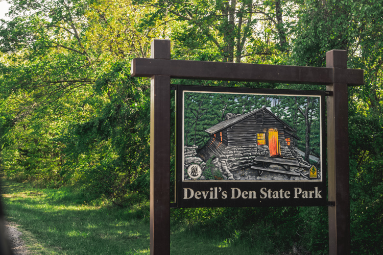 Sign for Devil's Den State Park, featuring an illustration of a cabin surrounded by trees, with green foliage in the background. The sign includes the park's name prominently displayed at the bottom.