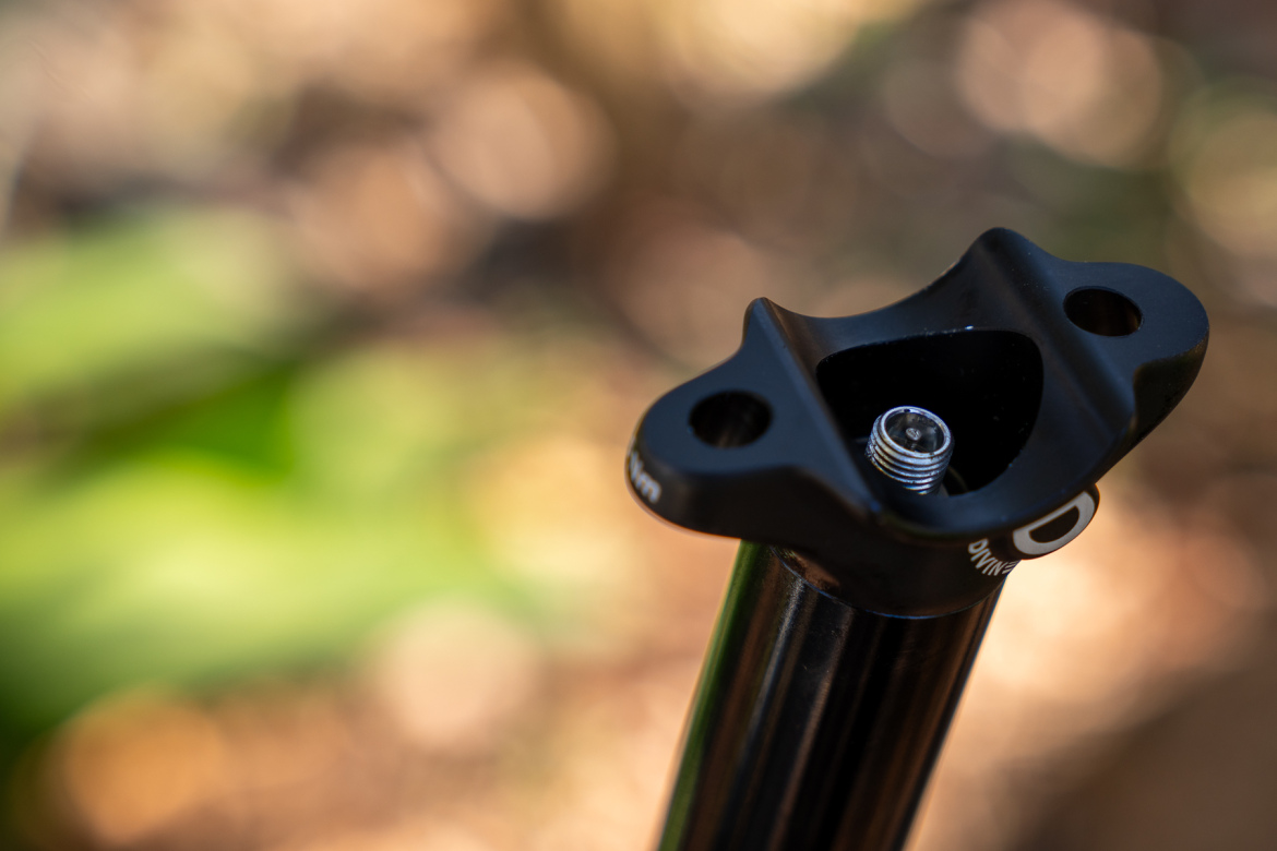 Close-up of a black bicycle seat post with a visible mounting interface, set against a blurred natural background. The focus is on the top of the seat post, showcasing its sleek design and threading for attachment.