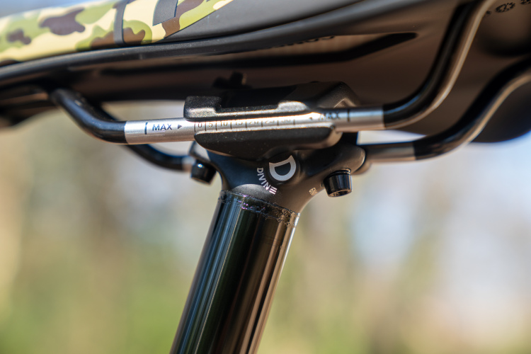 Close-up image of a bicycle seat post featuring a black and silver adjustment mechanism. The top of the post shows markings for maximum height and measurement indicators, with a visible brand logo on the attachment point. The background is softly blurred, suggesting an outdoor setting.
