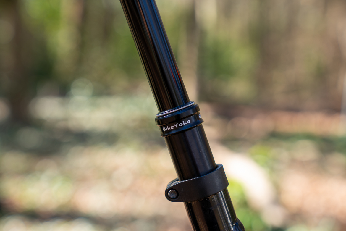 A close-up image of a black bike dropper post with a "BikeYoke" logo, featuring a metal collar and a quick-release lever, set against a blurred natural background.