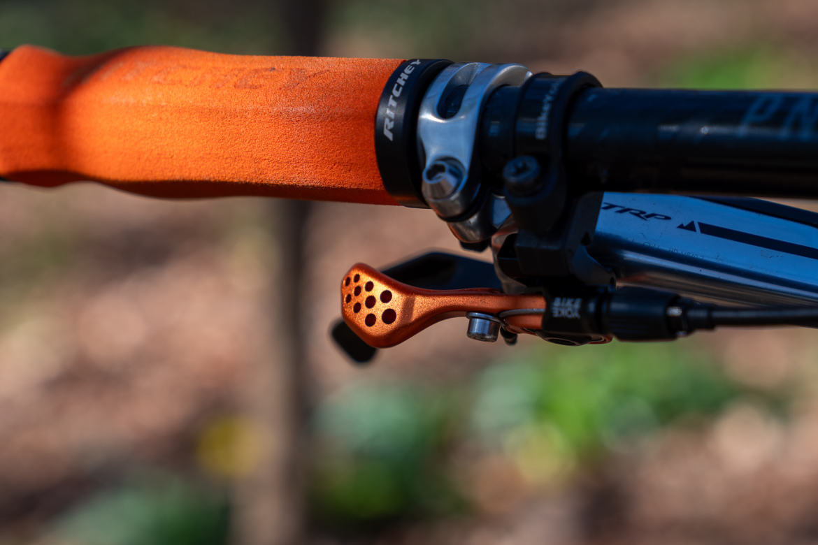 Close-up image of a bicycle handlebar featuring an orange grip and a metallic, perforated brake lever, with a blurred natural background.