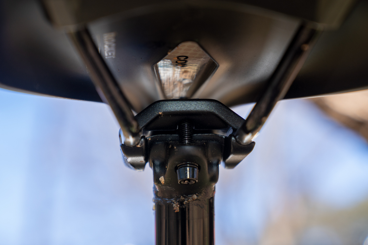 Close-up image of the underside of a black mount or bracket, showcasing a metal screw and hinge mechanism, with a blurred background of blue sky and trees.
