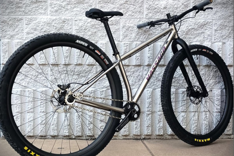 A sleek, modern mountain bike with a polished aluminum frame, wide black tires, and disc brakes, positioned against a textured white brick wall. The bike features a single chainring, flat handlebars, and Maxxis tires, reflecting its rugged design for off-road cycling.