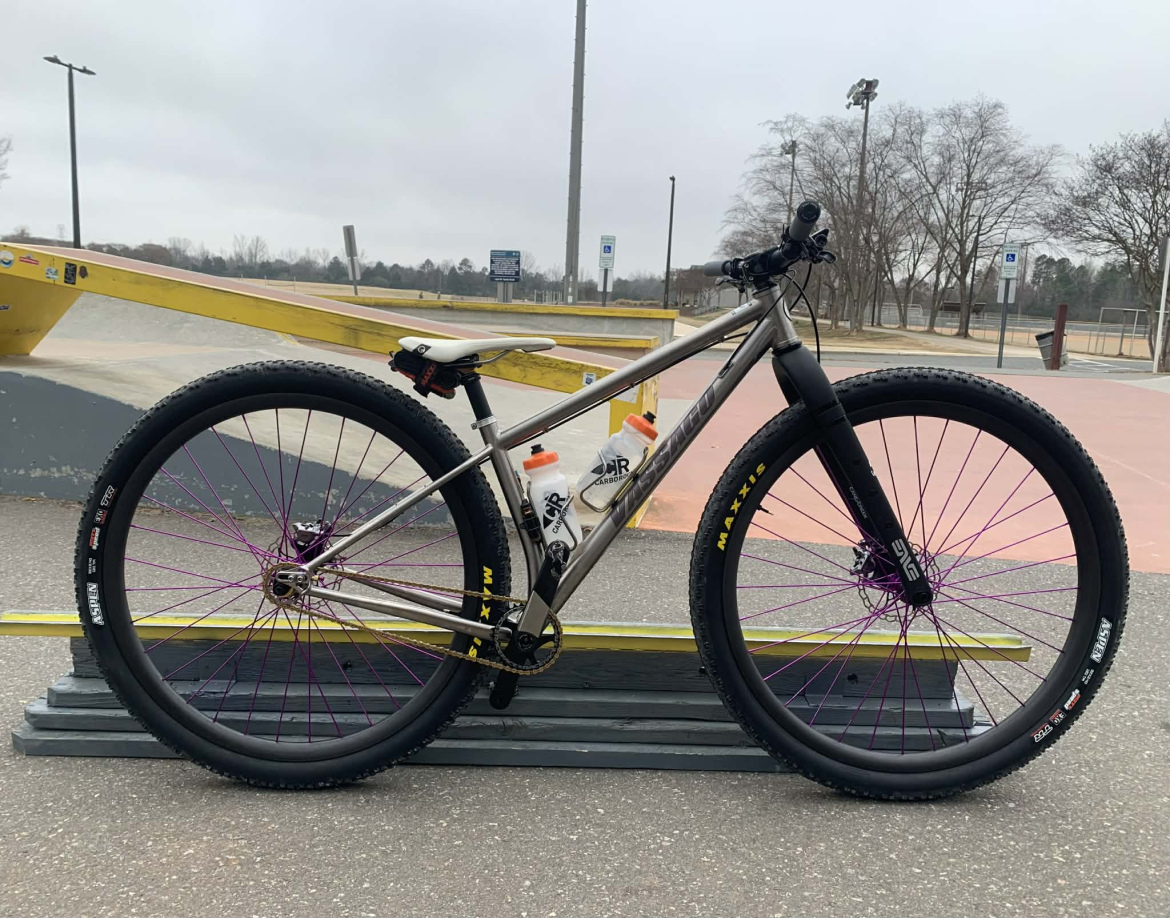 A custom mountain bike with a silver frame, purple spokes, and black tires. The bike is positioned on a wooden ramp at a skate park, with a water bottle mounted on the frame. The background includes trees and a cloudy sky, indicating an outdoor setting.